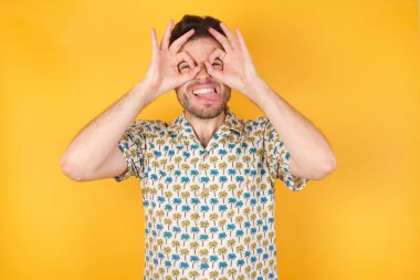 Yakışıklı genç adam dürbün gibi jest yapıyor. Dilini dışarı çıkarıyor, gözleri parmaklarından bakıyor. Çılgın ifade.