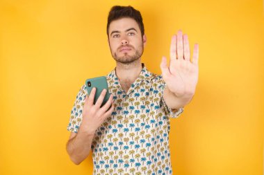 Sarı stüdyo arka planında akıllı telefonla poz veren yakışıklı genç adam.