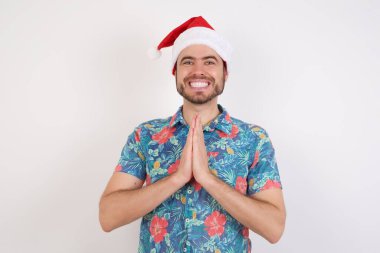 Noel Baba şapkalı genç adam elele dua ediyor af diliyor kendinden emin bir şekilde gülümsüyor..