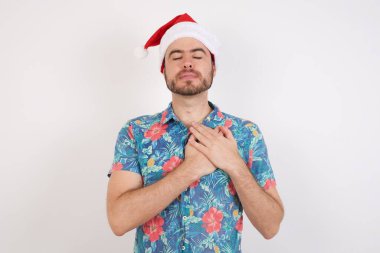 Noel Baba şapkalı genç adam elleri göğsünde gözleri kapalı gülümsüyor ve yüzünde minnet ifadesi var. Sağlık kavramı.