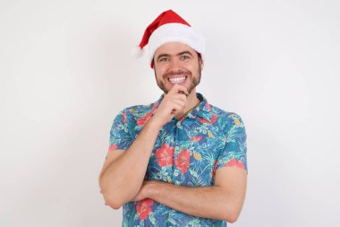 Noel Baba şapkalı genç adam ellerini kısmen çapraz yapar ve elleri çenesinin altında tutar, kameraya zevkle bakar. Mutlu duygu konsepti.