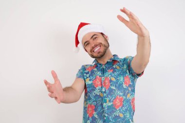 Noel Baba şapkalı genç adam kameraya bakıyor, sarılmak için kollarını açmış gülümsüyor. Mutluluğu kucaklayan neşeli bir ifade..