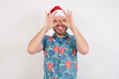 Noel Baba şapkalı genç adam dürbün gibi jest yapıyor. Dürbün gibi dilini çıkarıyor, gözler parmaklarından bakıyor. Çılgın ifade.