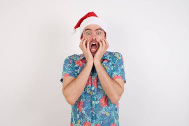 Noel Baba şapkalı genç adam heyecanını ve heyecanını ifade eder, çenesini indirir, elleri yanaklarında, gözleri dışarı fırlar