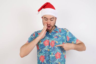 Şok olmuş Noel Baba şapkalı genç adam stüdyo arka planında poz veriyor. Şaşırtıcı bir şekilde aşağı bakıyor, işaret parmağıyla gömleğe bakıyor..