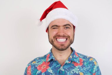 Noel Baba şapkalı genç adam mutlu bir şekilde gülümsüyor, kameraya neşeli bir şekilde göz kırpıyor, seninle flört ediyor. İnsan yüz ifadeleri ve duyguları