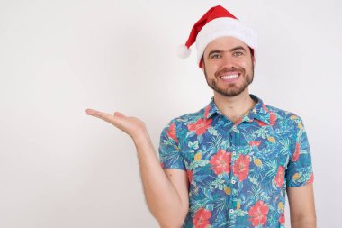 Noel Baba şapkalı genç adam, kameraya el ile bakıyor ve sunuyor..