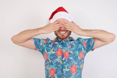 Noel Baba şapkalı genç adam elleri ile gözlerini kapatıyor neşeli ve komik gülümsüyor. Kör kavram.