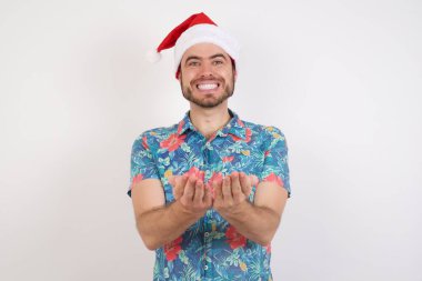 Noel Baba şapkalı genç adam avuçlarıyla gülümsüyor ve jest yapıyor. Tutun ve koruyun