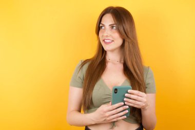 Sarı stüdyo arka planında modern akıllı telefonla poz veren güzel genç bir kadın.