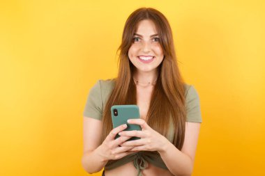 Sarı stüdyo arka planında modern akıllı telefonla poz veren güzel genç bir kadın.