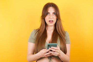 Sarı stüdyo arka planında modern akıllı telefonla poz veren güzel genç bir kadın.