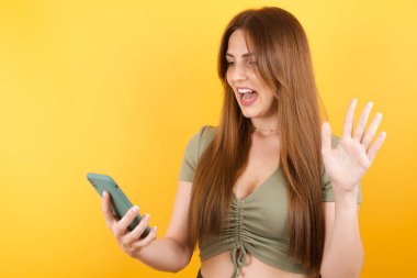 Sarı stüdyo arka planında modern akıllı telefonla poz veren güzel genç bir kadın.