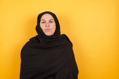 Başörtüsü takan olgun bir kadın dudaklarını büzüyor, kameraya yeşil gözlü bakıyor, kız arkadaşıyla eğleniyor, insan yüz ifadeleri takınıyor.