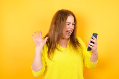 Güzel, olgun bir kadın sarı stüdyo arka planında akıllı telefonuyla poz veriyor.