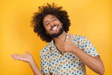 Yakışıklı genç adam avucunu gösteriyor ve baş parmağıyla hareket ediyor, mutlu ve neşeli gülümsüyor..