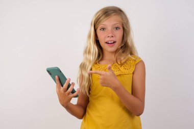 Stüdyo duvarında akıllı telefon pozu veren küçük tatlı bir kız fotoğrafı.