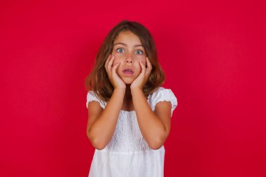 Güzel mavi gözlü küçük beyaz kız heyecanını ve heyecanını ifade eder, çenesini indirir, elleri yanaklarında, gözleri fırlar