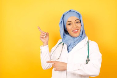 Kadın doktor işaret parmağı boşluğuyla en düşük fiyatları gösteriyor. İzole edilmiş sarı arkaplan giyer.