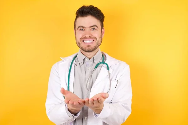 隔離された黄色の背景の上に立っている医学的な均一な聴診器を身に着けている若い白人医師の男性手のひらで一緒に笑顔を受信またはジェスチャーを与える ホールドと保護 — ストック写真
