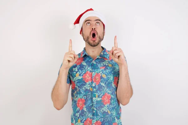 Mladý Muž Klobouku Santa Užaslý Překvapený Vzhlíží Ukazuje Prsty Zvednutými — Stock fotografie