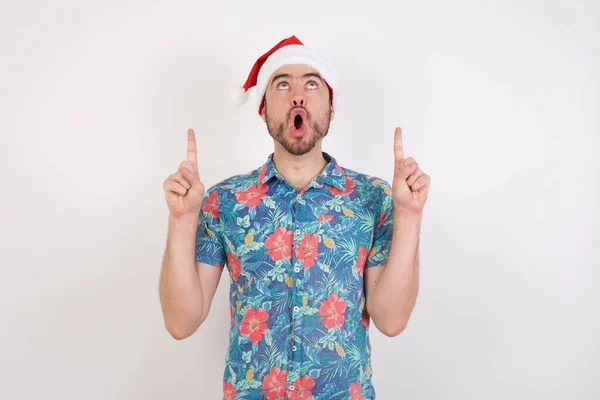 Mladý Muž Klobouku Santa Užaslý Překvapený Vzhlíží Ukazuje Prsty Zvednutými — Stock fotografie