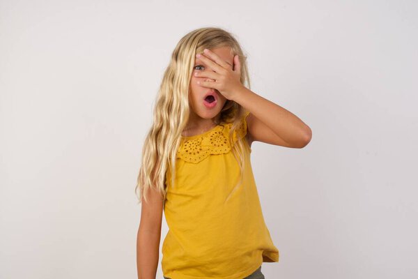 cute little girl peeking in shock covering face and eyes with hand, looking through fingers with embarrassed expression.