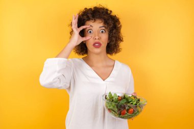 Stüdyoda kıvırcık saçlı, sarı duvarlı beyaz bir kadın. Elinde taze salata ile cam bir kase tutuyor. Parmaklarıyla gözlerini açıyor. 