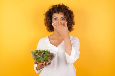 Stüdyoda kıvırcık saçlı, sarı duvarlı beyaz bir kadın. Elinde taze salata yiyeceği olan cam bir kase tutuyor. Ağzını eliyle kapatıyor. 
