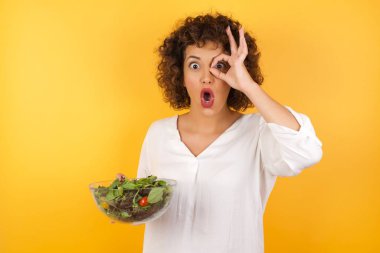 Beauitul 'lu kadın elinde salatayla bir kase tutuyor. Şaşırmış yüz ifadesiyle şaşırmış, parmaklarının arasından bakan gözler. İnançsız ifade.