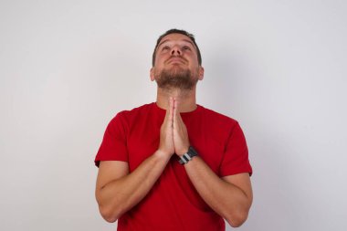 Kısa saçlı, kırmızı tişörtlü, izole edilmiş gri arka planda duran bir adam ellerini birleştirip af diliyor. 