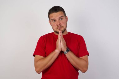 Kısa saçlı, kırmızı tişörtlü, izole edilmiş gri arka planda duran bir adam ellerini birleştirip af diliyor. 