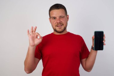 Elde cep telefonu tutan kırmızı tişörtlü yakışıklı beyaz adam, tamam işareti parmaklarıyla gösteriyor. 