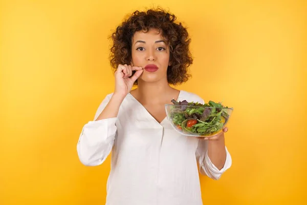 Young woman with salad  standing over isolated yellow background mouth and lips shut as zip with fingers. Secret and silent, taboo talking.