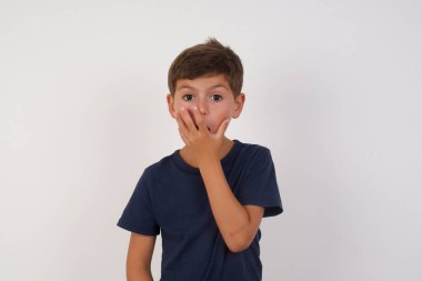 İzole edilmiş beyaz arka plan üzerinde duran günlük tişört giyen duygusal, güzel bir çocuk şaşkınlıktan nefesi kesiliyor, ağzı avuç içi ile kapanıyor, kameraya şok olmuş gibi bakıyor. Tepki konsepti.