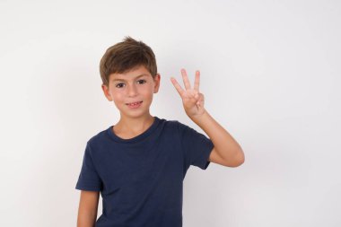 İzole edilmiş beyaz arka plan üzerinde duran, kendinden emin ve mutlu bir şekilde gülümseyen, üç numaralı parmaklarıyla işaret eden, sıradan tişört giyen güzel çocuk..