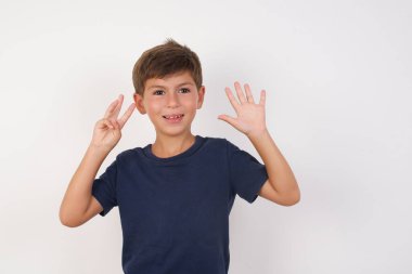 Güzel çocuk, izole edilmiş beyaz arka plan üzerinde sıradan bir tişört giyer ve 8 numaralı parmaklarıyla gülümseyerek mutlu ve kendinden emin bir şekilde durur..