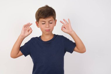 Güzel çocuk, izole edilmiş beyaz arka plan üzerinde sıradan bir tişört giyiyor, rahatlıyor ve parmaklarıyla meditasyon hareketi yaparken gözleri kapalı gülümsüyor. Yoga konsepti.