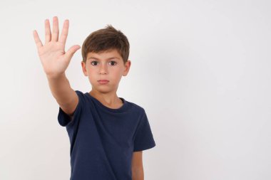 Güzel çocuk, izole edilmiş beyaz arka plan üzerinde sıradan bir tişört giyip, avuç içiyle el hareketi yapıyor. Yüzünde olumsuz ve ciddi bir jest olan uyarı ifadesi 