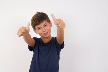 Güzel bir çocuk, izole edilmiş beyaz arka plan üzerinde duran günlük tişört giyiyor. Elle olumlu bir jest yapmayı onaylıyor. Gülümsüyor ve başarı için seviniyor. Kazanan jest.