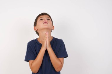 Güzel çocuk, izole edilmiş beyaz arka plan üzerinde sıradan bir tişört giyiyor, elleri bağlı yalvarıyor ve dua ediyor. Yüzünde umut ifadesi var. Çok duygusal ve endişeli. Soruyorum.