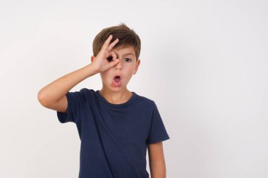 Güzel bir çocuk, izole edilmiş beyaz arka plan üzerinde sıradan bir tişört giyiyor. Şaşırmış bir yüz ve parmaklarından bakan bir gözle iyi bir hareket yapıyor. İnançsız ifade.
