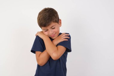 Güzel çocuk, izole edilmiş beyaz arka plan üzerinde sıradan bir tişört giyip mutlu ve pozitif bir şekilde kendine sarılıyor, kendinden emin gülümsüyor. Sevgi ve özgüven.