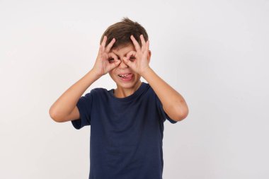 Güzel çocuk, izole edilmiş beyaz arka plan üzerinde sıradan bir tişört giyiyor, dürbün gibi jest yapıyor, gözleri parmaklarından bakıyor. Çılgın ifade.