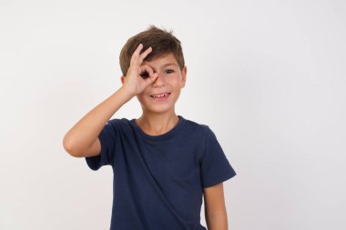 Güzel çocuk günlük tişört giyip izole edilmiş beyaz arka planda dikiliyor, el hareketleriyle gülümsüyor, gözleriyle mutlu yüzle parmaklarının arasından bakıyor..