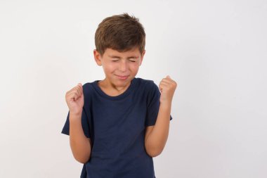 Güzel çocuk, izole edilmiş beyaz arka plan üzerinde sıradan bir tişört giyiyor, kollarını kaldırarak ve gözleri kapalı zaferi kutlayarak başarı için heyecanlanıyor. Kazanan konsept.