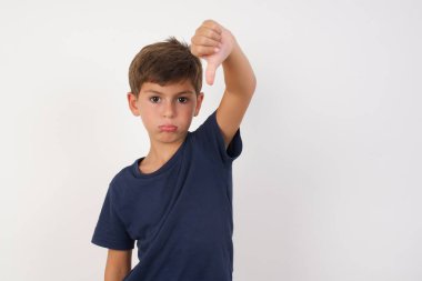 Güzel bir çocuk, izole edilmiş beyaz arka plan üzerinde duran, kızgın, sinirli, hayal kırıklığına uğramış ya da hoşnutsuz, ciddi bir bakışla baş parmaklarını gösteren bir tişört giyiyor.