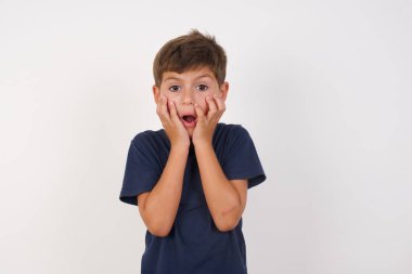 Sersemlemiş, sıradan tişörtler giyen, beyaz arka planda duran, heyecan ve heyecanı ifade eden, çenesini düşük, elleri yanaklarında, gözleri fırlayan güzel bir çocuk.