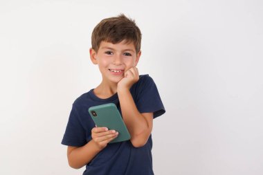 Neşeli, rahat bir tişört giyen, izole edilmiş beyaz arka plan üzerinde duran, cep telefonu cihazıyla poz veren, modern akıllı telefona mesaj yazan, boş zamanlarında komik videolar izleyen, iyi internet bağlantılarından hoşlanan güzel bir çocuk., 