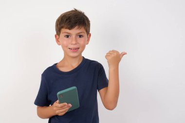 Güzel çocuk rahat bir tişört giyiyor, beyaz arka plan üzerinde duruyor, akıllı telefonla mesaj atıyor ve gülümseyen bir gülümsemeyle baş parmağıyla onu işaret ediyor.
